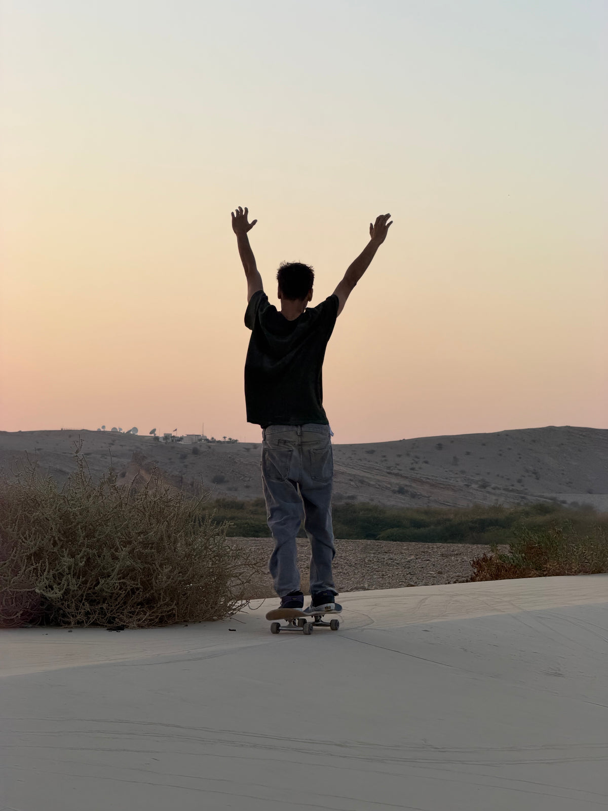 Oman: a skater raises his arms on his board at dusk in the desert. Photo by Mike Boisvert / The Skate Nomad